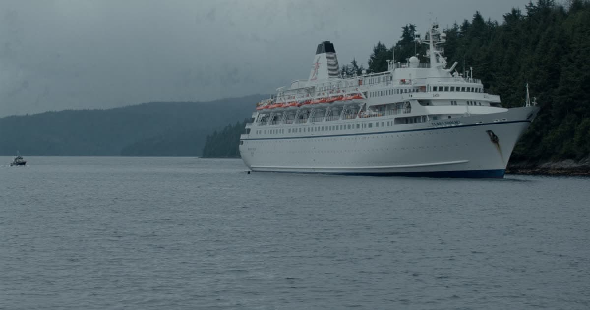 A Holland America style mid-size ship at anchor beside a forested coastline