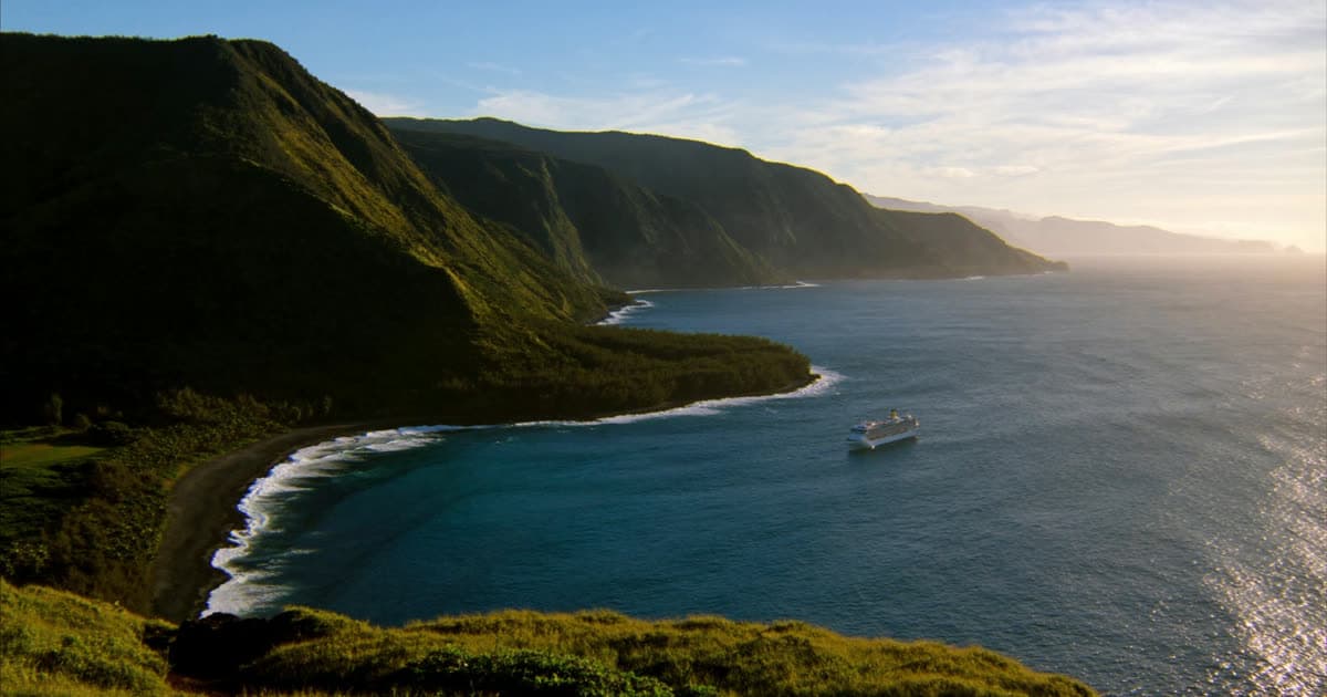 A cruise ship passing a volcanic Hawaiian coastline with green cliffs